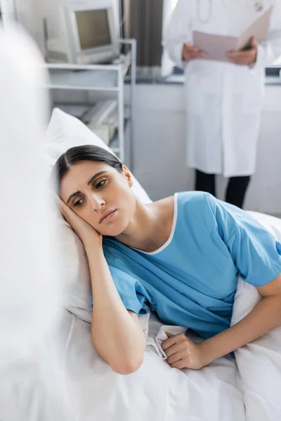 Displeased woman in patient gown lying on bed near blurred doctor in hospital ward — Stock Photo