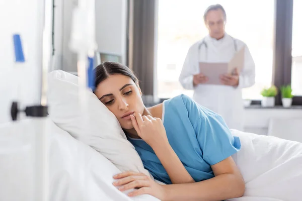 Brunette woman in patient gown lying on bed near blurred doctor in clinic - foto de stock