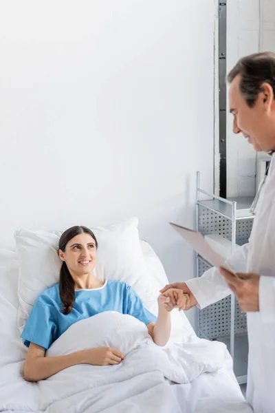 Улыбающийся пациент держит за руку пожилого врача с бумажной папкой в клинике — стоковое фото