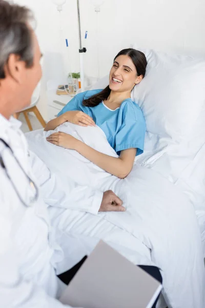 Веселый пациент, глядя на размытого старшего врача, лежащего на кровати в больничном отделении — стоковое фото