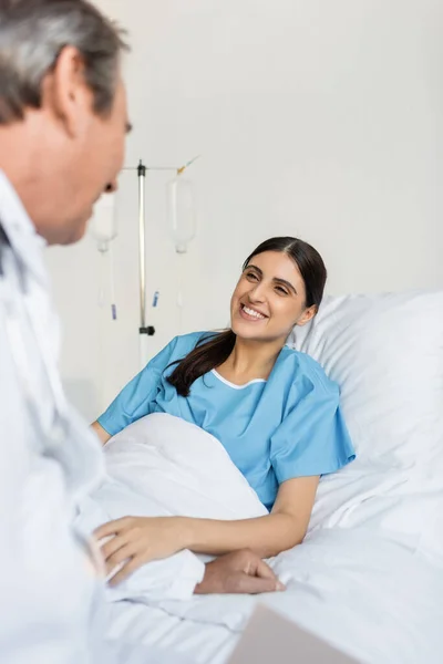 Smiling patient in gown talking to blurred doctor in hospital — Stock Photo