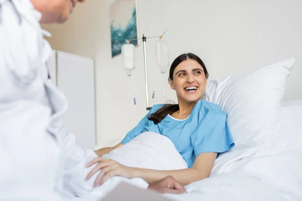 Paciente alegre em vestido deitado na cama perto de médico desfocado na clínica — Fotografia de Stock