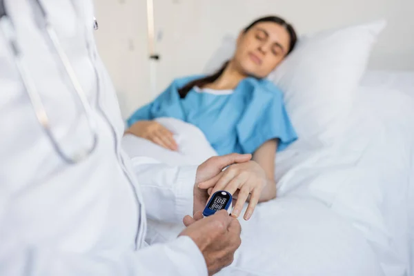 Elderly doctor wearing pulse oximeter on hand of blurred patient in clinic - foto de stock