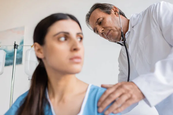 Senior doctor with stethoscope checking back of blurred patient in clinic — Stock Photo