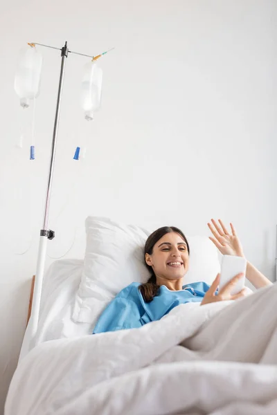Улыбающийся пациент с видеозвонком на смартфон рядом с внутривенной терапией в больнице — стоковое фото