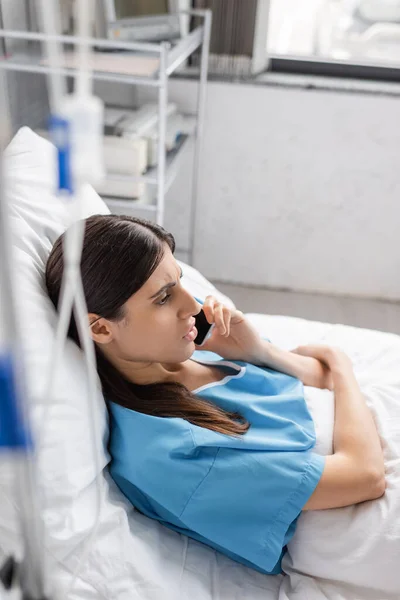 Пациент разговаривает на смартфоне во время лежания на кровати вблизи размытой внутривенной терапии в больнице — стоковое фото