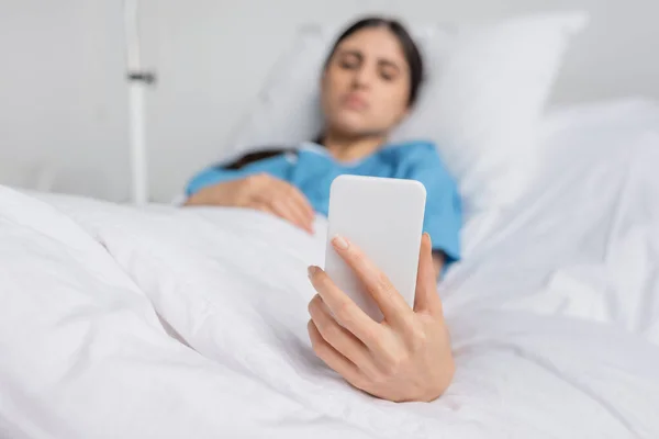 Blurred woman in patient gown using smartphone on bed in clinic - foto de stock