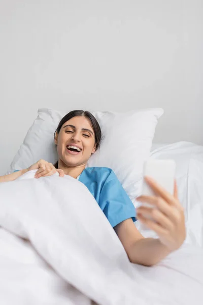 Positive woman in patient gown using cellphone on bed in hospital ward — Stock Photo