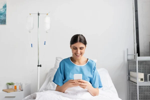 Happy woman in patient gown smiling while using smartphone on bed in clinic — Stock Photo