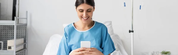 Paziente allegro utilizzando smartphone sul letto in clinica, banner — Foto stock