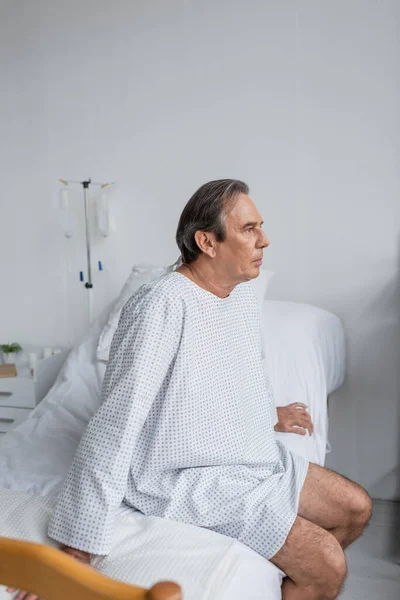 Седой пациент в халате сидит на кровати в больнице — стоковое фото