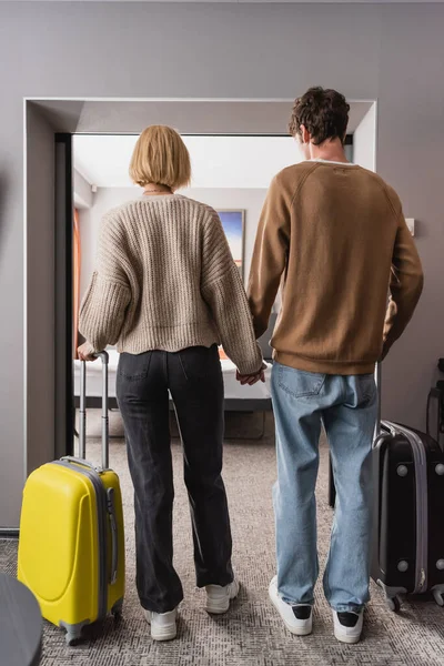 Вид ззаду молода пара з туристичними сумками, що стоять в готельних апартаментах і тримаються за руки — стокове фото