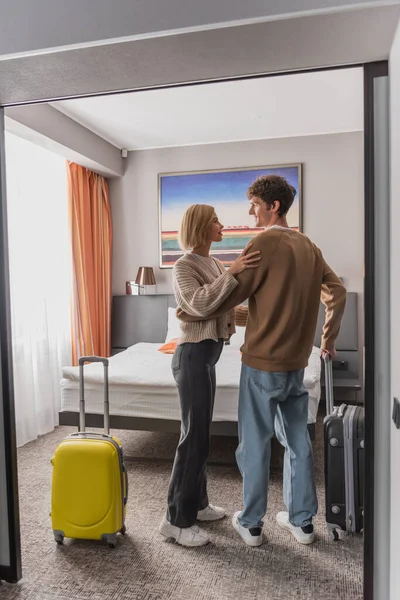Full length of happy young couple looking at each other near baggage in hotel bedroom - foto de stock