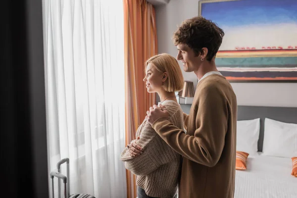 Side view of smiling man hugging girlfriend standing with crossed arms near window in hotel room — Stock Photo