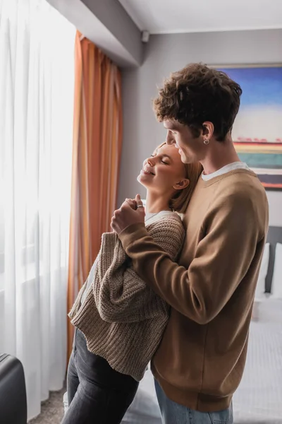 Happy blonde woman with closed eyes holding hands with smiling boyfriend in hotel apartments — Stock Photo