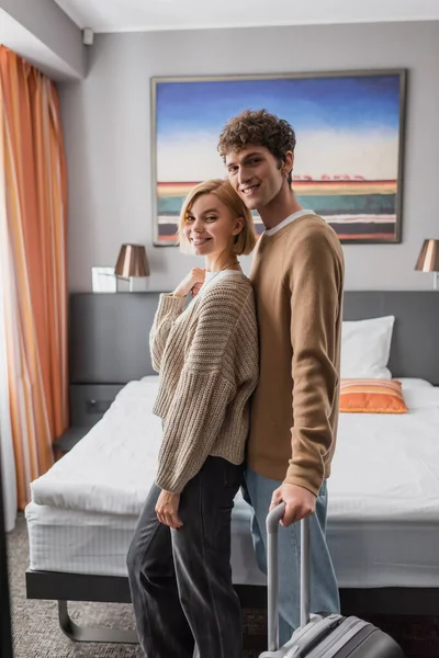 Веселые молодые путешественники улыбаются перед камерой, стоя с чемоданом в спальне отеля — стоковое фото