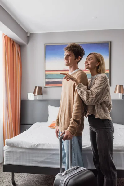 Happy man with travel bag near blonde woman pointing with finger in hotel room — Stock Photo