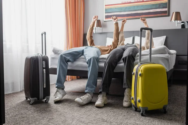 Young couple of tourists showing yeah gesture on bed near suitcases in modern hotel bedroom — Photo de stock