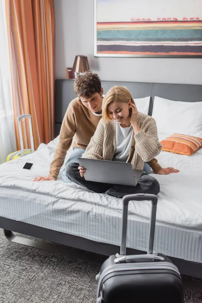 Smiling woman sitting on hotel bed with laptop near young boyfriend and suitcase on foreground - foto de stock
