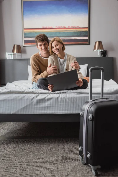 Веселая пара путешественников, смотрящих в камеру, сидя на кровати с ноутбуком возле чемодана в гостиничном номере — стоковое фото