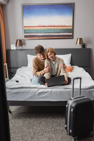 Happy woman sitting with laptop near smiling boyfriend on bed in modern hotel - foto de stock