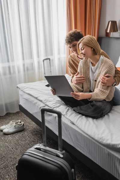 Молодой человек обнимает подругу сидя со скрещенными ногами и ноутбуком на кровати в апартаментах отеля — стоковое фото