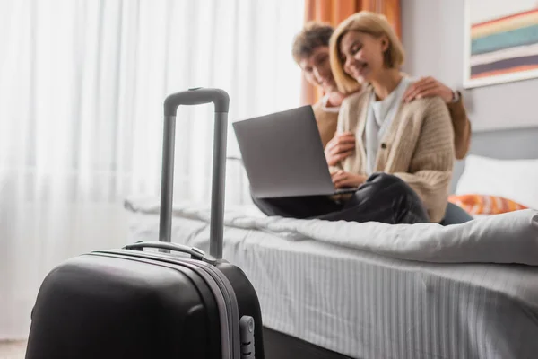 Selective focus of suitcase near happy couple watching movie on laptop on hotel bed on blurred background - foto de stock
