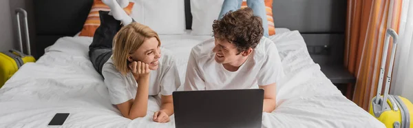 Cheerful young couple looking at each other near laptop and smartphone on bed in hotel suite, banner - foto de stock