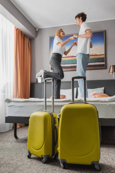 Веселая молодая пара, держась за руки, стоя на кровати возле чемоданов в отеле — стоковое фото