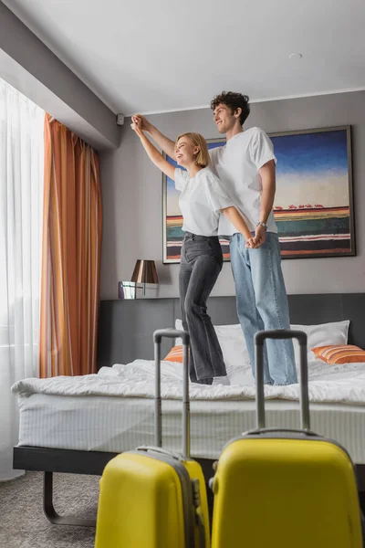 Full length of joyful couple standing on bed and holding hands while having fun in hotel room near travel bags — Photo de stock