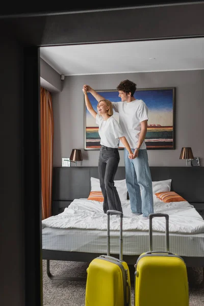 Couple of young travelers holding hands while having fun on hotel bed near luggage on foreground — Stockfoto