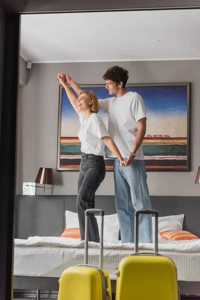 Young and cheerful couple holding hands and having fun while standing on bed in modern hotel apartments — Photo de stock