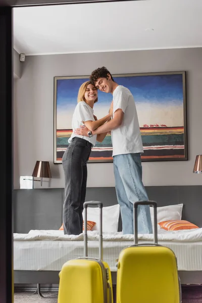 Cheerful couple standing on bed and embracing near suitcases in modern hotel suite — Fotografia de Stock