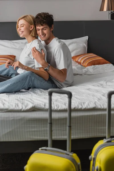 Довольный мужчина обнимает подругу, сидя на кровати рядом с чемоданами на размытом переднем плане — стоковое фото