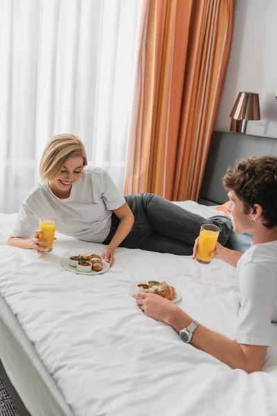 Молодые путешественники завтракают с вкусными круассанами и апельсиновым соком на кровати в отеле — стоковое фото