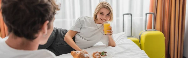 Happy woman with glass of orange juice looking at blurred boyfriend on bed in hotel apartments, banner - foto de stock