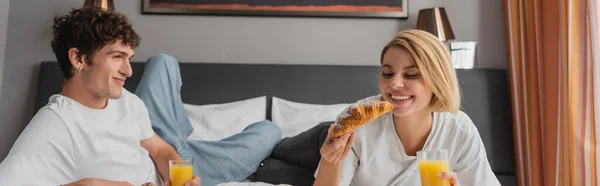 Happy blonde woman holding delicious croissant and orange juice near smiling boyfriend in hotel bedroom, banner — Stockfoto