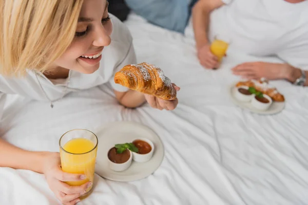 Happy blonde woman holding tasty croissant and orange juice while having breakfast on hotel bed near blurred boyfriend — Stock Photo