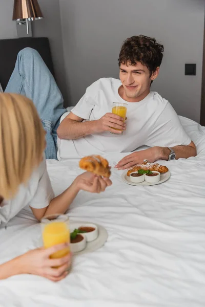 Cheerful man holding orange juice and looking at blonde girlfriend having breakfast on hotel bed — Stock Photo