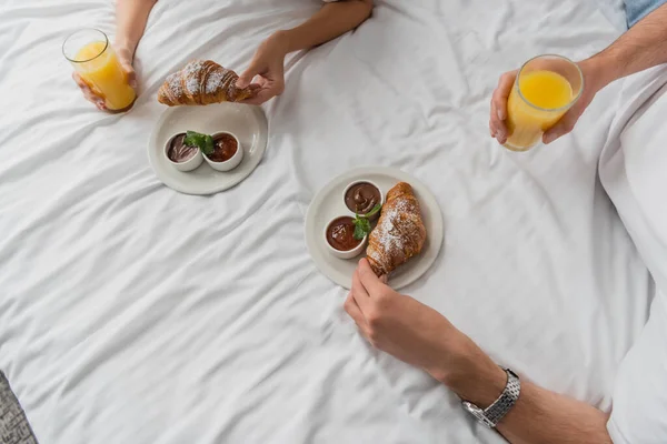 Top view of cropped couple holding orange juice near delicious croissants and jam with chocolate paste on hotel bed — Stock Photo