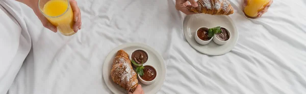 Top view of cropped couple with glasses of orange juice near croissants with chocolate paste and jam on bed in hotel, banner - foto de stock