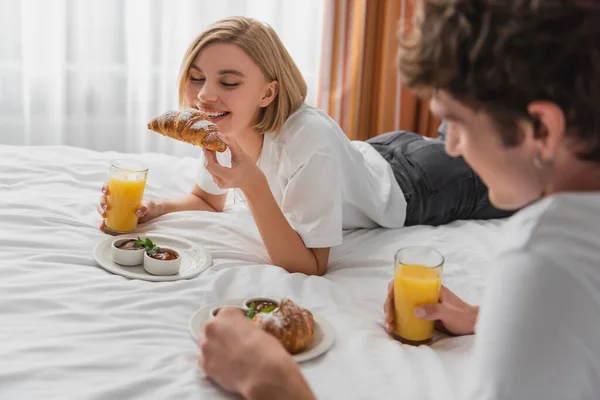 Happy woman with orange juice eating tasty croissant near blurred boyfriend on bed in hotel apartments — Stock Photo
