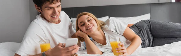 Smiling man using smartphone near happy girlfriend with glass of orange juice on bed in hotel, banner — Stockfoto