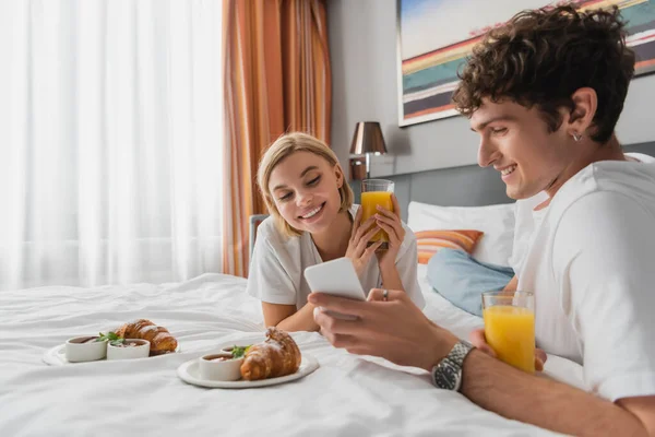 Young man showing mobile phone to smiling girlfriend near croissants and orange juice on hotel bed — Stock Photo