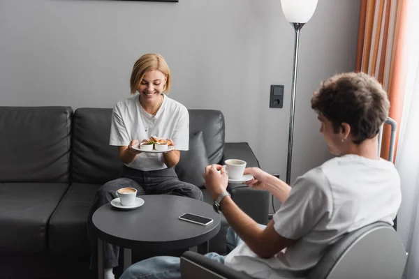 Щаслива жінка тримає смачний сніданок біля хлопця з чашкою і смартфоном на кавовому столі в готельному номері — стокове фото