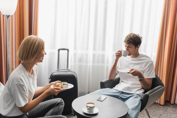 Young man drinking coffee near smiling girlfriend with croissant in modern hotel room — Stockfoto