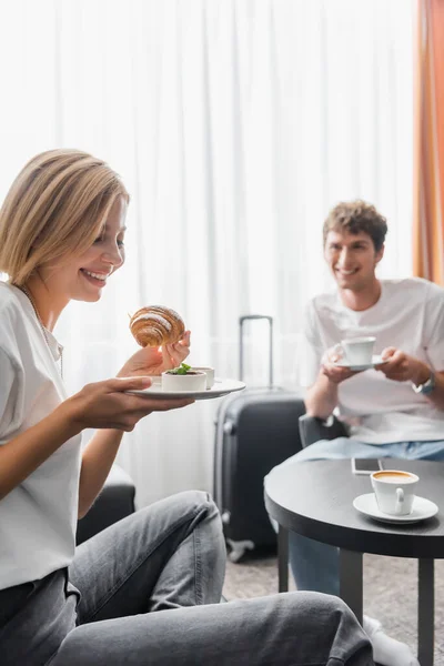 Веселая женщина держит вкусный круассан рядом с парнем с чашкой кофе в номере отеля на размытом фоне — стоковое фото
