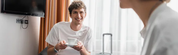 Happy young man holding coffee cup near girlfriend in hotel room on blurred foreground, banner — Photo de stock
