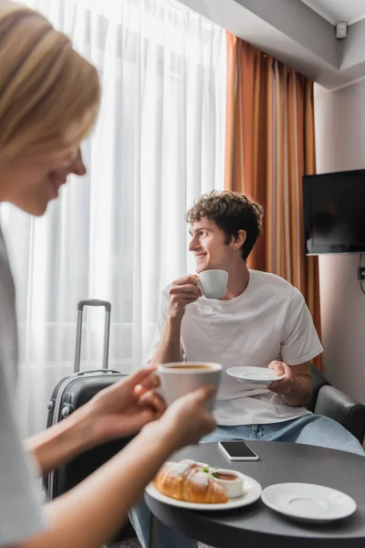 Smiling man sitting in hotel room with coffee cup and looking away near girlfriend on blurred foreground — Stockfoto