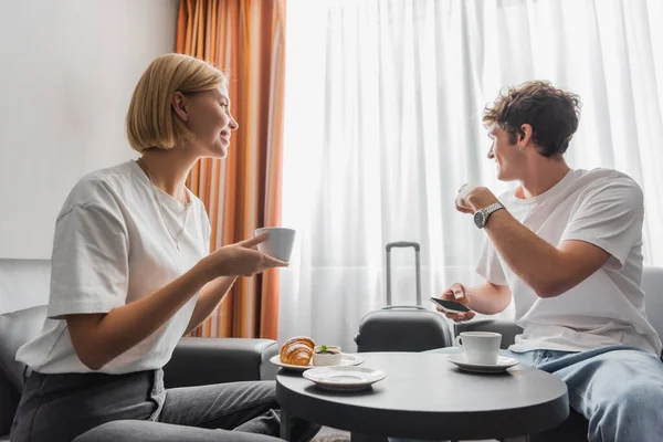 Young man holding smartphone and pointing at window near girlfriend with coffee cup in hotel room — Stockfoto